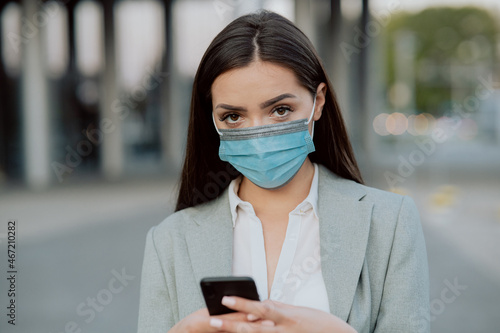 A beautiful woman dressed in a shirt and jacket with protective mask on face is standing in front of corporate building, break from work, holding a phone in hands, answering messages, email