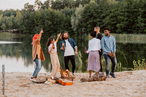 Students friends of different nationalities skin colors meet on beach by lake light bonfire fry bread sausages play guitar music dance, have fun, drink beer from bottles which they hold in their hands