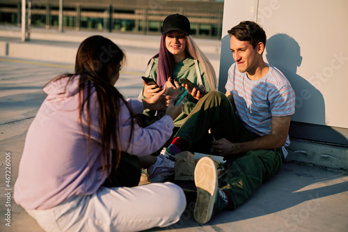 Happy teenage friends are sitting in urban exterior and using their phones for hanging on social media.