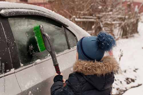 a child with a brush in his hands and in winter clothes cleans the car from snow