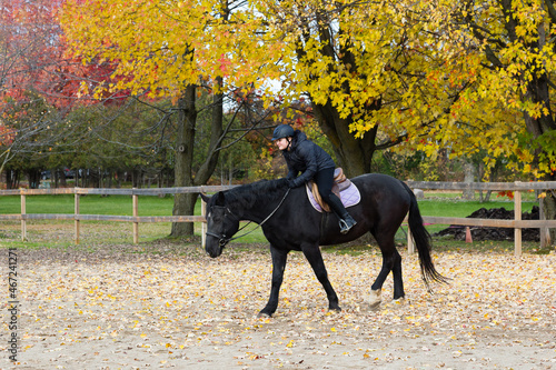 Horizontal shot of cute pre-teen girl trotting on black horse in exterior arena during a fall afternoon