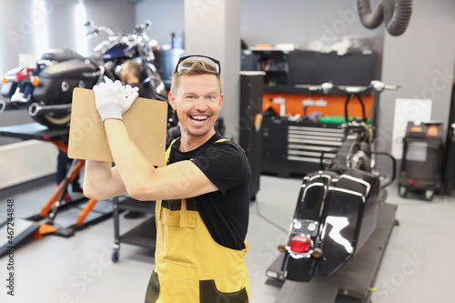 Happy male repairman holding clipboard with documents near motorcycle in car repair shop