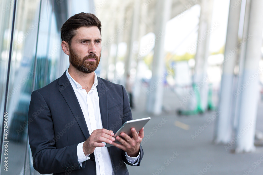 stylish bearded businessman in formal business suit standing working with tablet in hands on background modern office building outside. Man using smartphone or uses mobile phone outdoors city street stylish bearded businessman in formal business suit standing working with tablet in hands on background modern office building outside. Man using smartphone or uses mobile phone outdoors city street