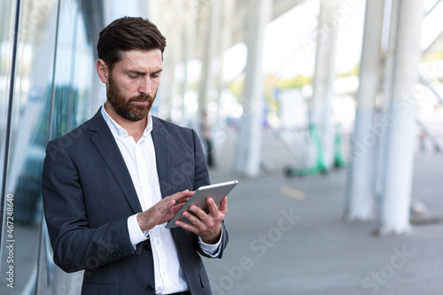 stylish bearded businessman in formal business suit standing working with tablet in hands on background modern office building outside. Man using smartphone or uses mobile phone outdoors city street