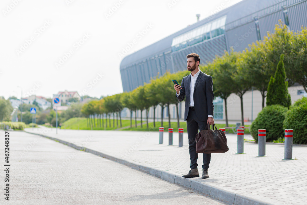 stylish bearded business man with travel bag and mobile phone in hands walks on city street modern urban background. Businessman in a suit goes and uses a smartphone. tourist trip. caucasian manager stylish bearded business man with travel bag and mobile phone in hands walks on city street modern urban background. Businessman in a suit goes and uses a smartphone. tourist trip. caucasian manager