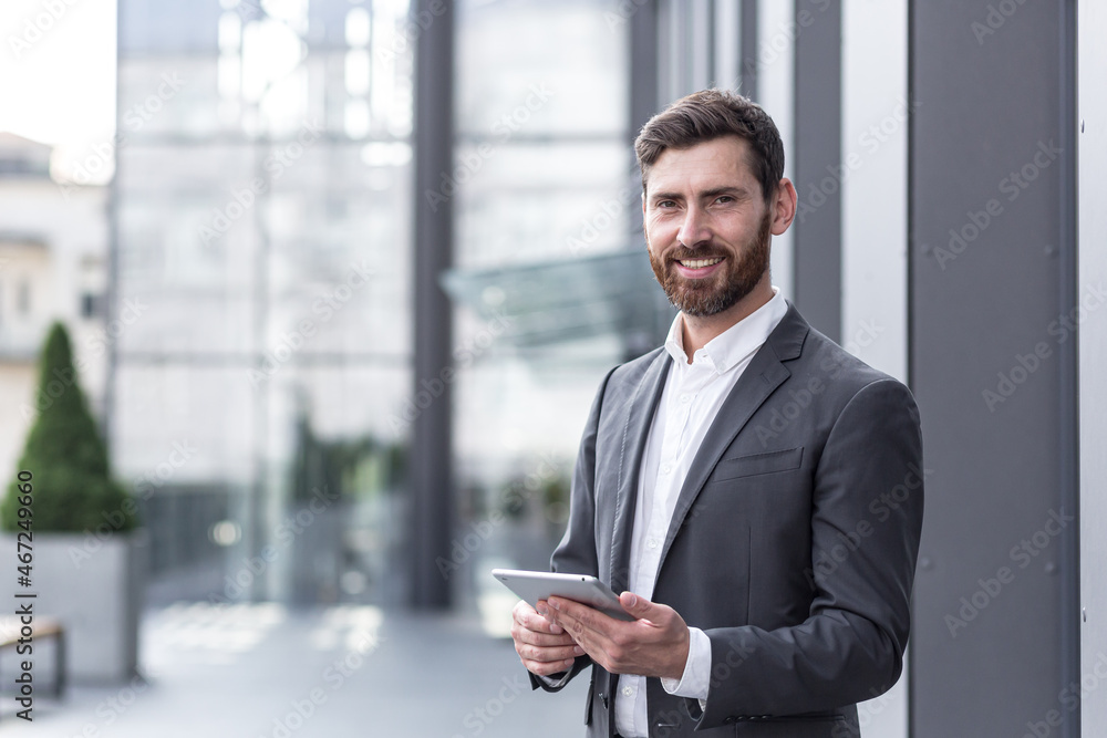 Fototapeta premium Cheerful and successful businessman works outdoors near the office holds a tablet, smiles reads good news