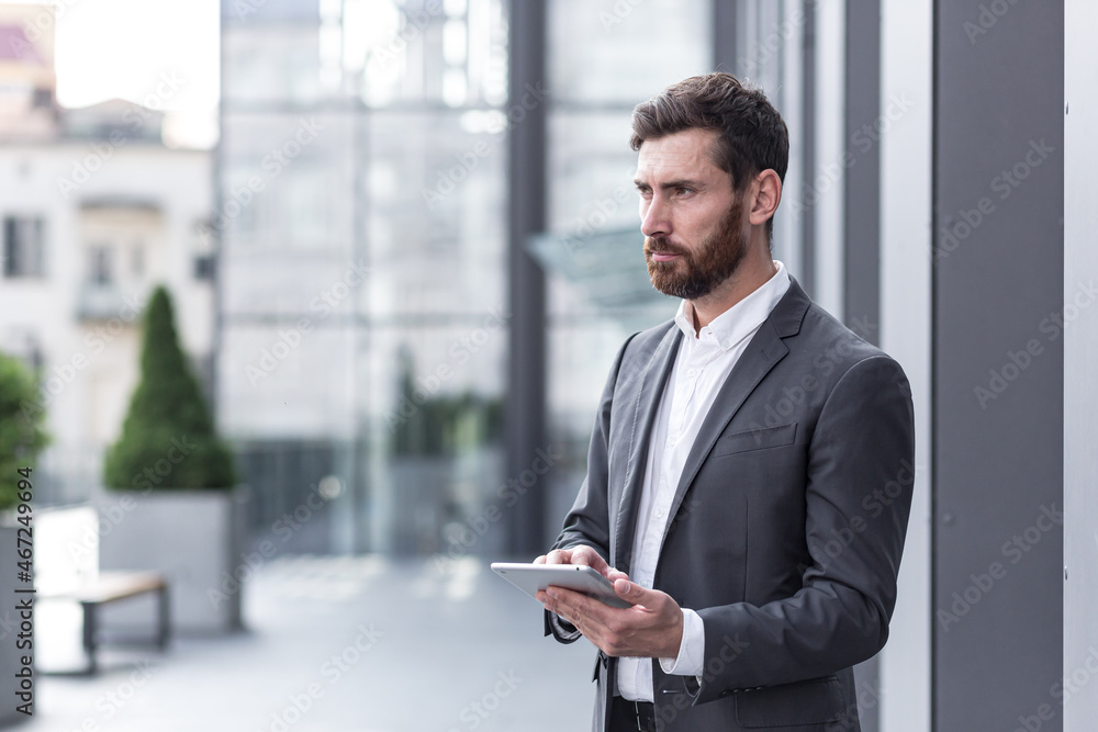 stylish bearded businessman in formal business suit standing working with tablet in hands on background modern office building outside. stylish bearded businessman in formal business suit standing working with tablet in hands on background modern office building outside.