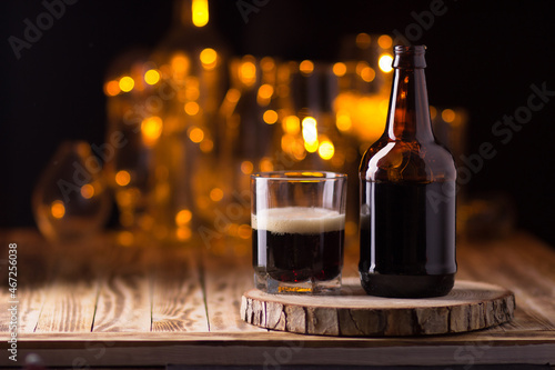 An open bottle of beer and a half-filled glass on the table on a dark background with bright yellow bokeh lights.