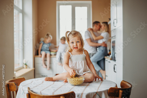 A little girl is sitting on the table at home in the morning and eating a ready breakfast. In the background is a happy young family.
