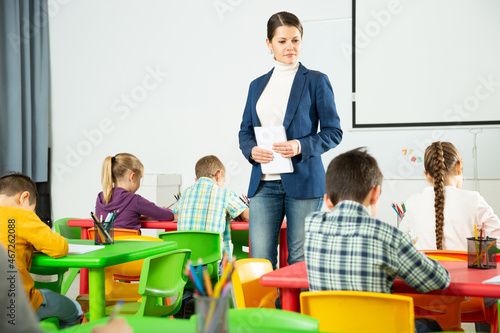 Young woman teacher moving among pupils desks, controlling writing of test work by elementary school students in lesson..