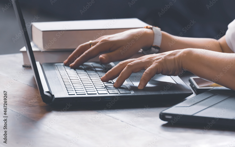 Closeup of woman hands typing on laptop keyboard with books and various devices on wooden table in home office room Closeup of woman hands typing on laptop keyboard with books and various devices on wooden table in home office room