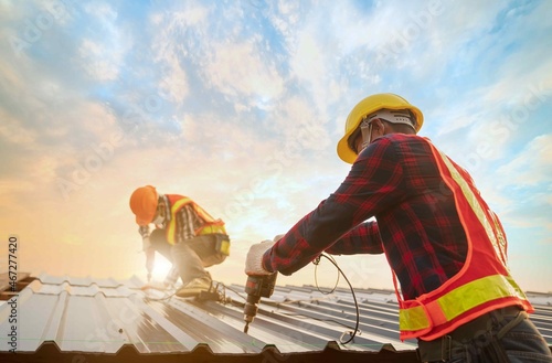 Foto Roofer worker in protective uniform wear gloves, using electric screw drill installing iron roof or metal sheet on top of the new roof,Concept of residential building safty under construction