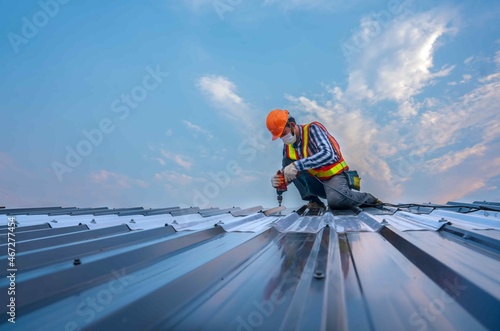 Roofer worker in protective uniform wear gloves, using electric screw drill installing iron roof or metal sheet on top of the new roof,Concept of residential building safty under construction.