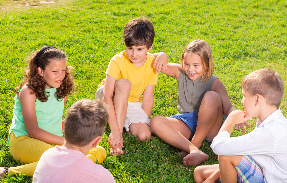 Fototapeta premium Young boys and girls sitting on green grass and discussing something funny.