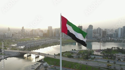 The Flag of the United Arab Emirates waving in the air, the Blue sky and city development in the Background, The national symbol of UAE over Sharjah's Flag Island. 4k Footage photo