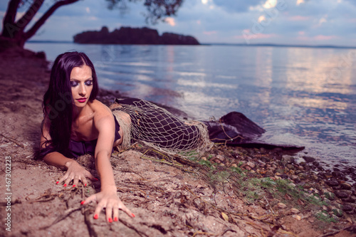 Seducing Mermaid Posing With Net Near Sea on Rocks While Wearing Artistic Makeup and Black Shiny Tail Covered With Colorful Spangles Posing as Queen of Sea photo