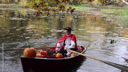 young man dressed as gondolier in gondola boat decorated with pumpkins on pond in autumn park celebrates Halloween and having fun, concept of Halloween carnival or costume party