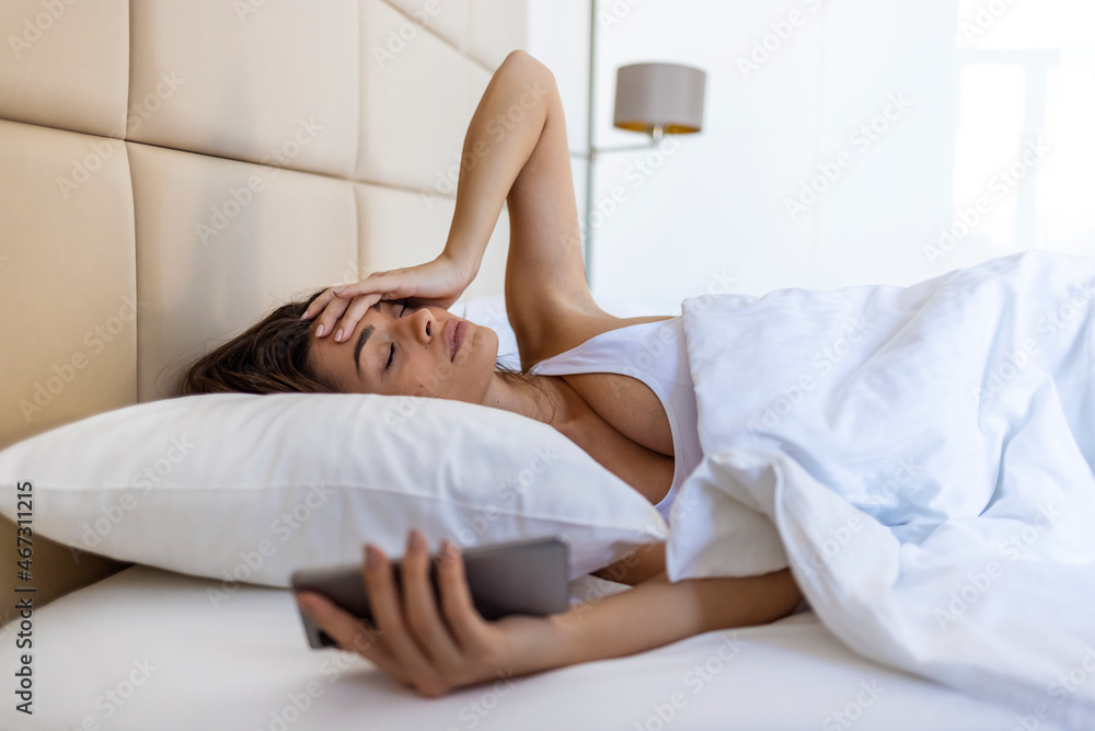 Fototapeta premium Woman lying in bed reaching to turn off the alarm on her mobile phone in morning. Sleepy woman being woken up by smartphone alarm. Young woman waking up in bed and checking her smartphone