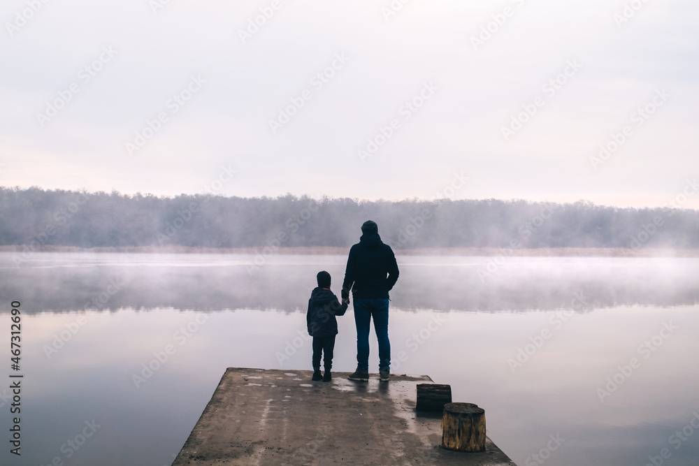 Dad and son on a mountain lake in fog. Magic morning. The fog spreads beautifully over the water surface. The trees are almost invisible. Clouds and sky reflect off the surface.
