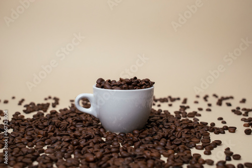 White Cup  full of coffee beans. With a lots of beans around. Light brown background.