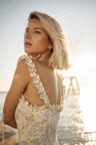 Gorgeous caucasian young woman posing tenderly at sunrise near sea. Beauty with bob haircut stands half sideways in sundress and looks into distance embracing herself. 