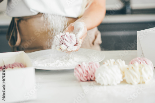 Skilled female confectioner covers delicious homemade multicolored marshmallows with powdered sugar in plate at table in light kitchen