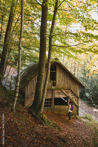 young backpacker woman observing an abandoned wooden house in the middle of the forest