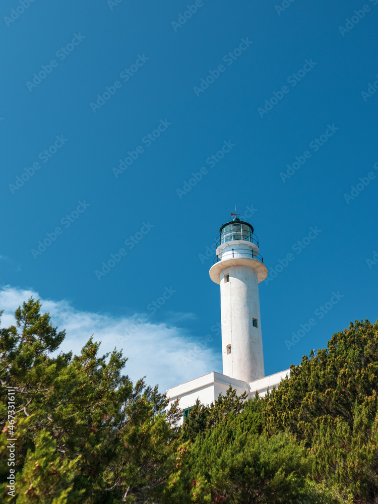 White lighthouse tall tower in pine bushes greenery on vivid blue sky in Greece, Ionian sea. Scenic travel destination. Lefkada island. Vertical White lighthouse tall tower in pine bushes greenery on vivid blue sky in Greece, Ionian sea. Scenic travel destination. Lefkada island. Vertical