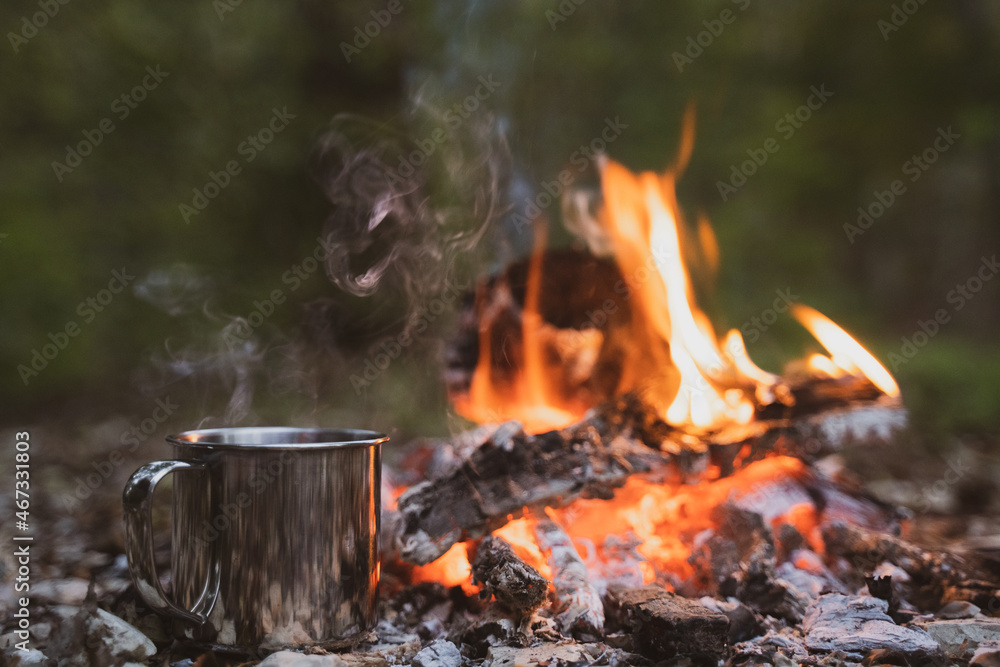 A mug with a hot drink is heated into a fire in a camp. Cozy rest, dusk A mug with a hot drink is heated into a fire in a camp. Cozy rest, dusk