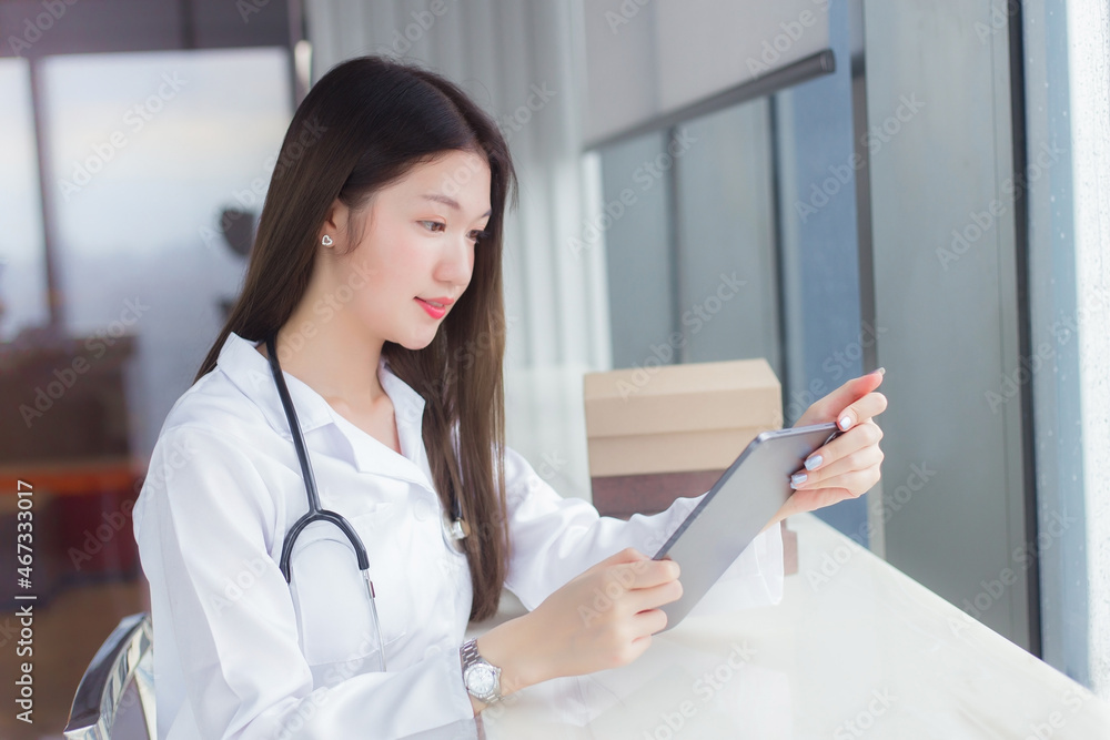Young Asian woman doctor dressed in white medical coat sits on chair at a hospital library to search some information on a tablet for treatment. Young Asian woman doctor dressed in white medical coat sits on chair at a hospital library to search some information on a tablet for treatment.