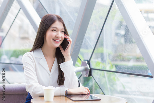 Beautiful young Asian professional business female in a white shirt is  while using your smartphone in your hand to talk she is sitting happy smile working at coffee shop.
