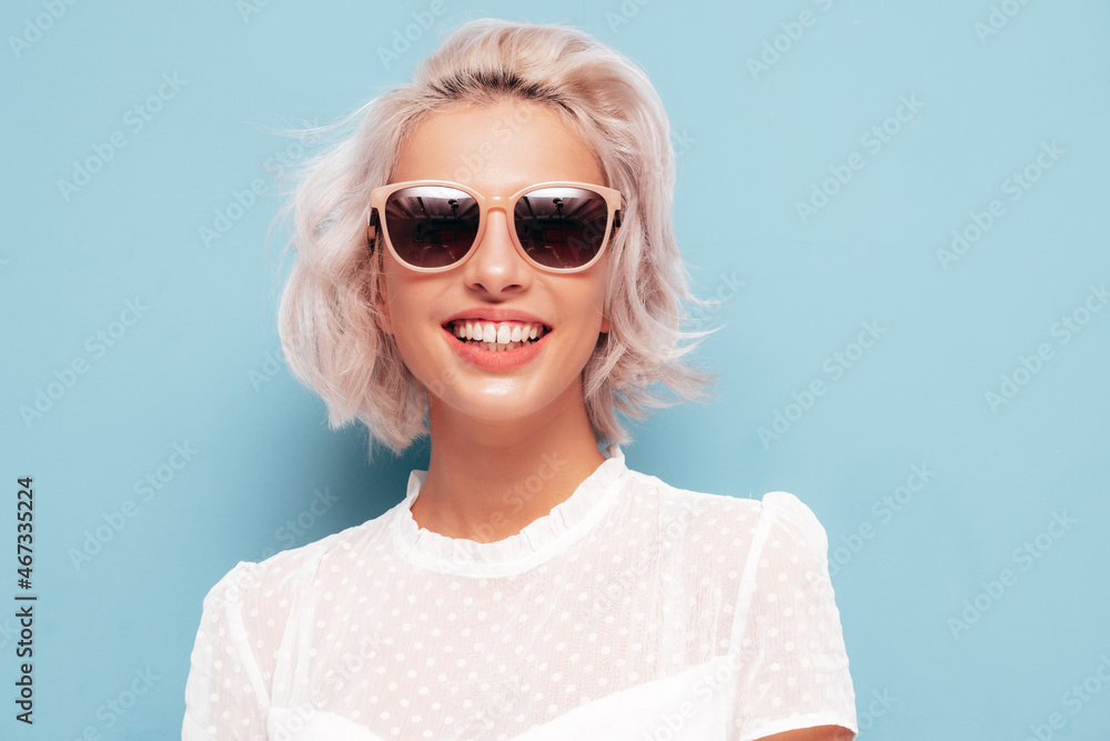Portrait of young beautiful smiling female in trendy summer white dress. Sexy carefree blond woman posing near blue wall in studio. Positive model having fun indoors. Cheerful and happy in sunglasses Portrait of young beautiful smiling female in trendy summer white dress. Sexy carefree blond woman posing near blue wall in studio. Positive model having fun indoors. Cheerful and happy in sunglasses