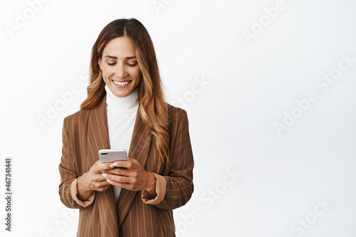 Smiling corporate woman order taxi in app, chatting in messanger app on smartphone, looking at her phone screen with pleased face, white background photo