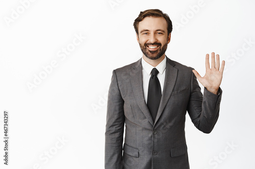 Business people. Handsome smiling businessman waving raised hand, saying hello, welcoming you, standing in grey suit over white background