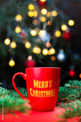 Morning coffee in a red cup on the red surface with Christmas tree lights and spruce branches in the background