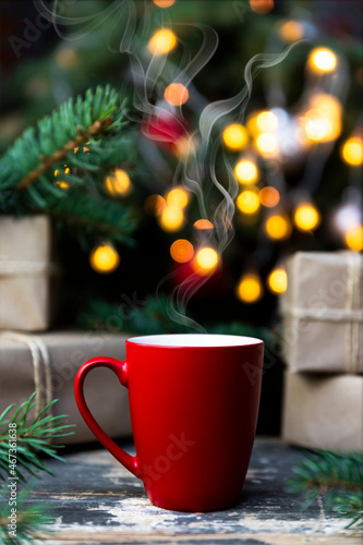 Coffee in a red cup on the rustic table with wrapped gifts, Christmas tree lights and spruce branches in the background