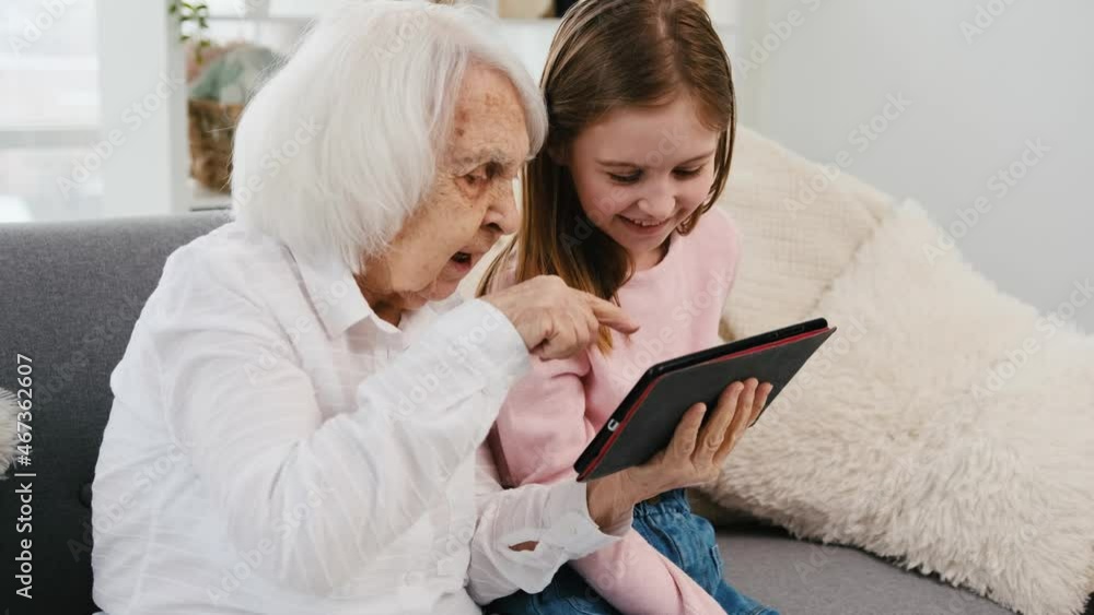 Granddaughter and grandmother sitting on the sofa with tablet and communicate. Concept of generations and technologies Granddaughter and grandmother sitting on the sofa with tablet and communicate. Concept of generations and technologies