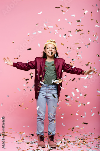 full length of cheerful preteen girl in stylish outfit smiling near falling confetti on pink.