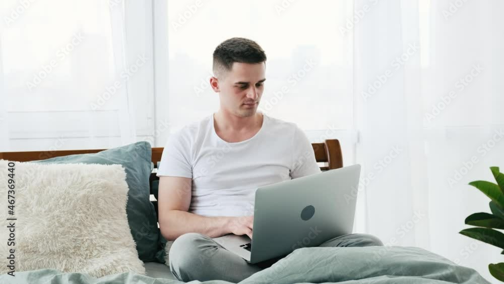 Young handsome man sitting on the bed with laptop in the sunny room. Guy wearing pajamas in his apartment during morning work from home. Man reading news from notebook staying in the bed relaxing Young handsome man sitting on the bed with laptop in the sunny room. Guy wearing pajamas in his apartment during morning work from home. Man reading news from notebook staying in the bed relaxing