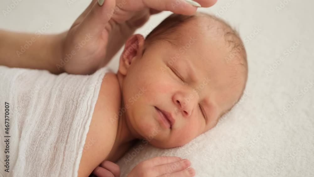 Closeup portrait of adorable newborn baby sleeping and mother hand strocking his head. Swaddled infant child in white fabric napping. Beautiful kid resting Closeup portrait of adorable newborn baby sleeping and mother hand strocking his head. Swaddled infant child in white fabric napping. Beautiful kid resting