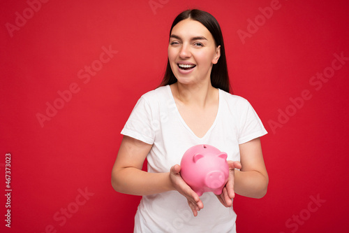 Portrait of happy positive smiling young beautiful attractive brunette woman with sincere emotions wearing casual white t-shirt isolated over red background with free space and holding pink piggy box © Ivan Traimak