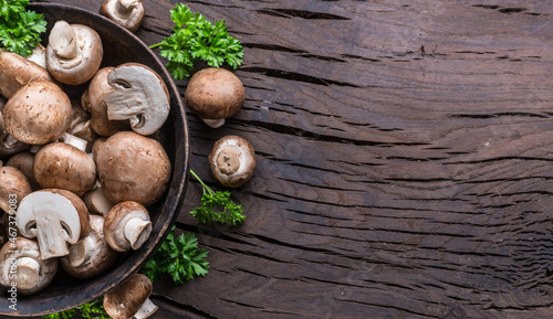 Brown colored common mushrooms in wooden bowl on wooden table with herbs. Top view.