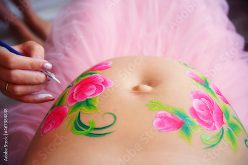 Close-up of a pregnant belly, on which the artist paints with a brush aqua makeup of pink flowers with green leaves. High quality photo