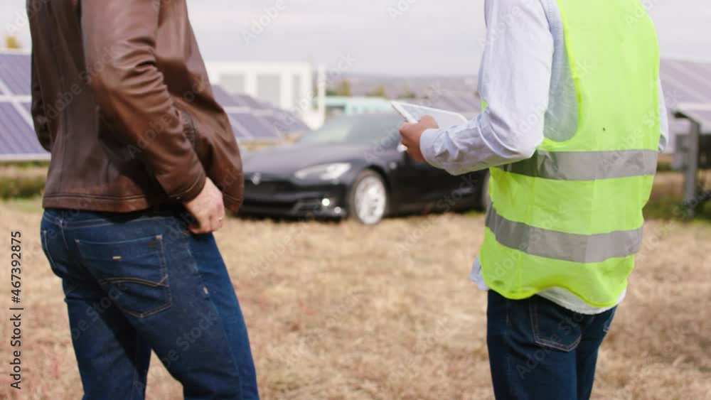 Details closeup taking video of ecological engineer and ecological worker together end the discussion at solar panels farm and continue to walking to the electric car beside of photovoltaic batteries Details closeup taking video of ecological engineer and ecological worker together end the discussion at solar panels farm and continue to walking to the electric car beside of photovoltaic batteries