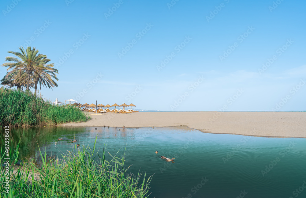 Paisaje de un lago verde con patos, una duna de arena de playa, y sombrillas y las aguas azules del  mar en el horizonte un día soleado con cielo azul de fondo.