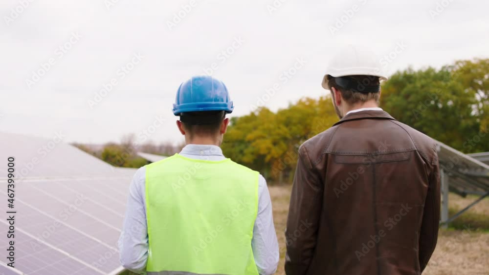 Green energy concept at solar panels farm taking video from the back of a two ecological workers walking through the photovoltaic batteries and discussing something they wearing safety helmets Green energy concept at solar panels farm taking video from the back of a two ecological workers walking through the photovoltaic batteries and discussing something they wearing safety helmets