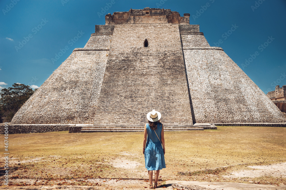 A young woman stands against the backdrop of the Mayan pyramid in Uxmal and enjoys the breathtaking view, from the back. Pyramid of in the ruins of Uxmal, Yucatan, Mexico,Unesco archaeological site - obrazy, fototapety, plakaty 