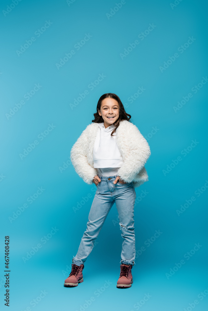 full length of happy preteen girl in faux fur jacket and winter boots standing with hands in pockets on blue. full length of happy preteen girl in faux fur jacket and winter boots standing with hands in pockets on blue.