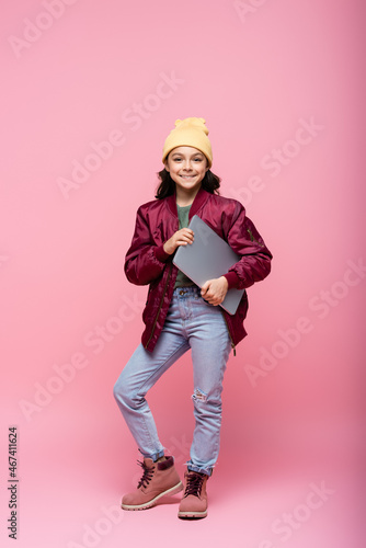 full length of stylish preteen girl in winter outfit posing with laptop and smiling on pink.