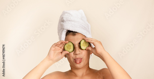 Portrait of a child on a spa day. Caucasian boy with towel on his head sticks out his tongue and puts cucumber slices in his eyes. Health and beauty spa day